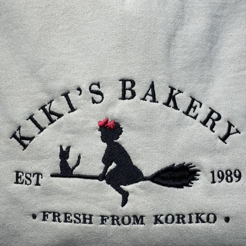 May include: White sweatshirt with an embroidered design. The design features the text "Kiki's Bakery" above a silhouette of a witch on a broom with a black cat. The text "EST 1989" and "FRESH FROM KORIKO" are also included.