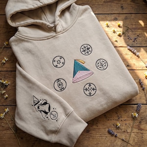 Op de afbeelding: Een beige hoodie met een geborduurd ontwerp op de borst en mouw. Op de borst staat een centrale kegelvormige hoed in groenblauw en roze, omringd door vijf zwarte cirkelvormige symbolen. De mouw is voorzien van extra zwarte geborduurde iconen.