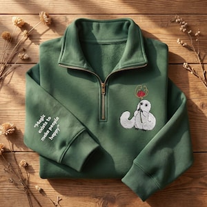 Op de afbeelding: Een bosgroene sweater met een goudkleurige kwartritssluiting. Op de borst staat een geborduurd wit wezen dat een rode bloem vasthoudt. De mouw is bedrukt met de witte tekst: "Magic exists to make people happy."