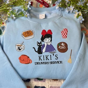 May include: Light blue sweatshirt with embroidered Kiki's Delivery Service design. Features Kiki, a black cat, and various food and delivery-related images. The text "KIKI'S DELIVERY SERVICE" is printed below the images.