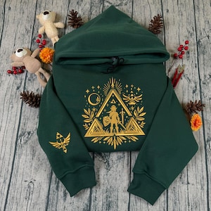 Peut inclure: Un sweat à capuche vert foncé avec un motif graphique doré sur le devant et la manche gauche. Le motif représente un triangle avec une figurine tenant une épée et un bouclier, entourée d'étoiles et d'autres symboles. Le sweat est en matière douce.