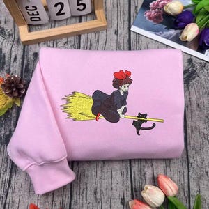 Puede incluir: Una sudadera rosa claro con un diseño bordado de una niña en una escoba con un gato negro. La niña lleva un vestido negro y un lazo rojo. El gato es negro. La sudadera está doblada sobre una superficie de madera.