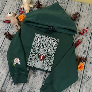 May include: A dark green hooded sweatshirt with a white and red graphic of a forest spirit on the front. The sweatshirt has a drawstring hood and an embroidered design on the left sleeve. The design features a forest spirit and leaves.