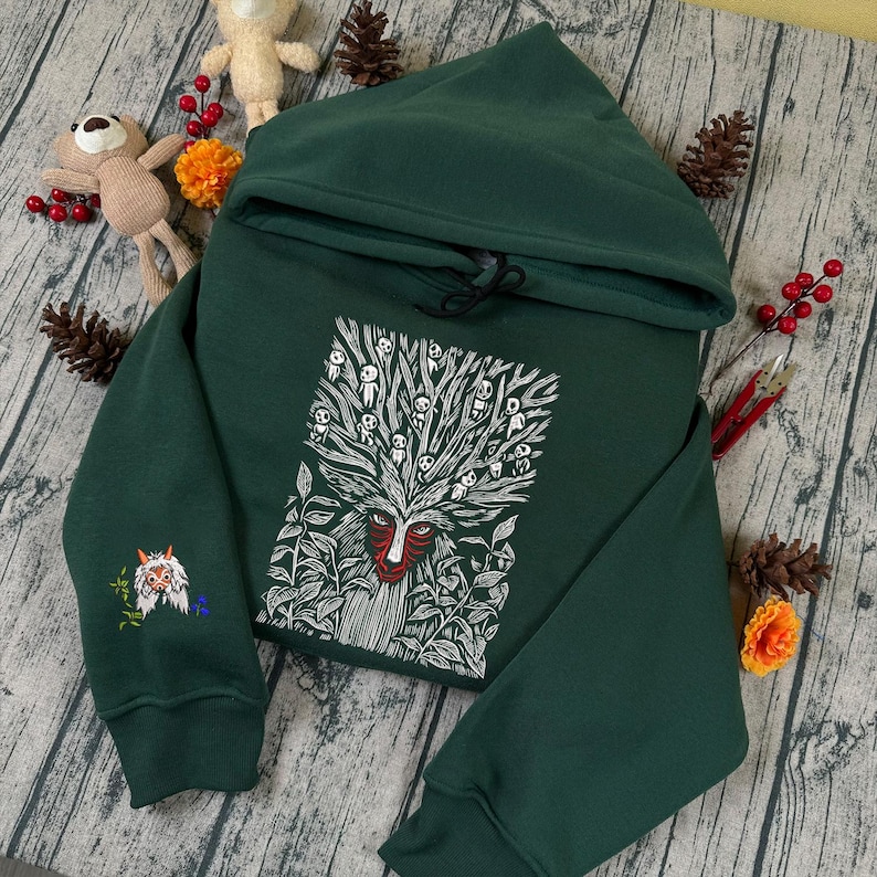 May include: A dark green hooded sweatshirt with a white and red graphic of a forest spirit on the front. The sweatshirt has a drawstring hood and an embroidered design on the left sleeve. The design features a forest spirit and leaves.