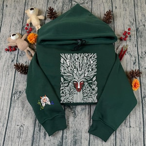 May include: A dark green hoodie with a hood and drawstring. The front features a white and red graphic of a forest spirit. The left sleeve has an embroidered design. The hoodie is displayed on a wooden surface with decorative elements.