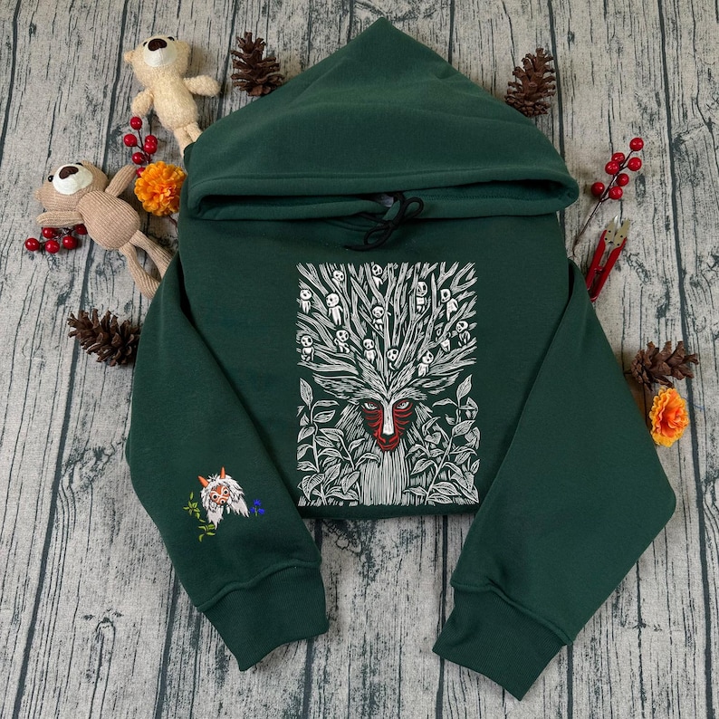 May include: A dark green hoodie with a hood and drawstring. The front features a white and red graphic of a forest spirit. The left sleeve has an embroidered design. The hoodie is displayed on a wooden surface with decorative elements.