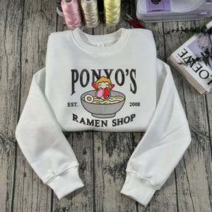 May include: White crew neck sweatshirt featuring the text "PONYO'S RAMEN SHOP" and a graphic of a ramen bowl. The design includes a cartoon character and the founding year, 2008. The sweatshirt has ribbed cuffs and hem.