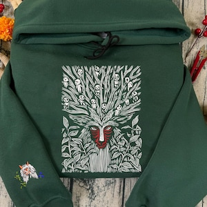 May include: A dark green hoodie featuring a white and red graphic of a forest spirit with a tree-like head and leaf details. The sleeve has an embroidered design. The hoodie has a drawstring and a hood.