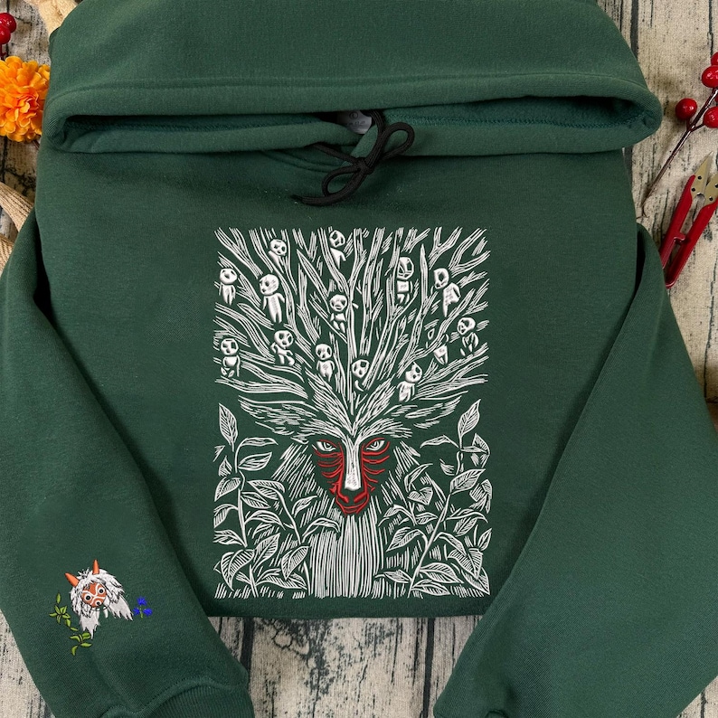 May include: A dark green hoodie featuring a white and red graphic of a forest spirit with a tree-like head and leaf details. The sleeve has an embroidered design. The hoodie has a drawstring and a hood.