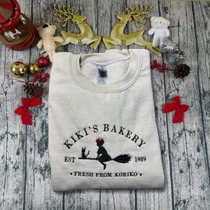 May include: Cream-colored sweatshirt with the embroidered text "Kiki's Bakery" featuring a witch on a broom, a black cat, and the year 1989. The phrase "Fresh from Koriko" is also embroidered on the sweatshirt.