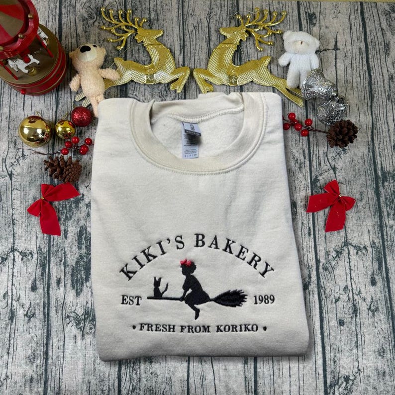 May include: Cream-colored sweatshirt with the embroidered text "Kiki's Bakery" featuring a witch on a broom, a black cat, and the year 1989. The phrase "Fresh from Koriko" is also embroidered on the sweatshirt.