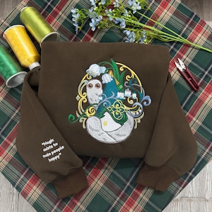 Könnte beinhalten: Ein dunkelbraunes Sweatshirt mit einer zentralen Stickerei einer weißen Kreatur mit großen Augen, umgeben von blau-weißen Maiglöckchen und goldenem Filigran. Der Ärmel enthält den gestickten Text "Magic exists to make people happy".