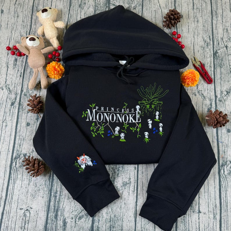 Pu&ograve; includere: Felpa con cappuccio nera con la scritta "Princess Mononoke" e un disegno ricamato di spiriti della foresta e un lupo. La felpa ha un cappuccio con coulisse e polsini a coste. Il design include accenti verdi, bianchi e blu.