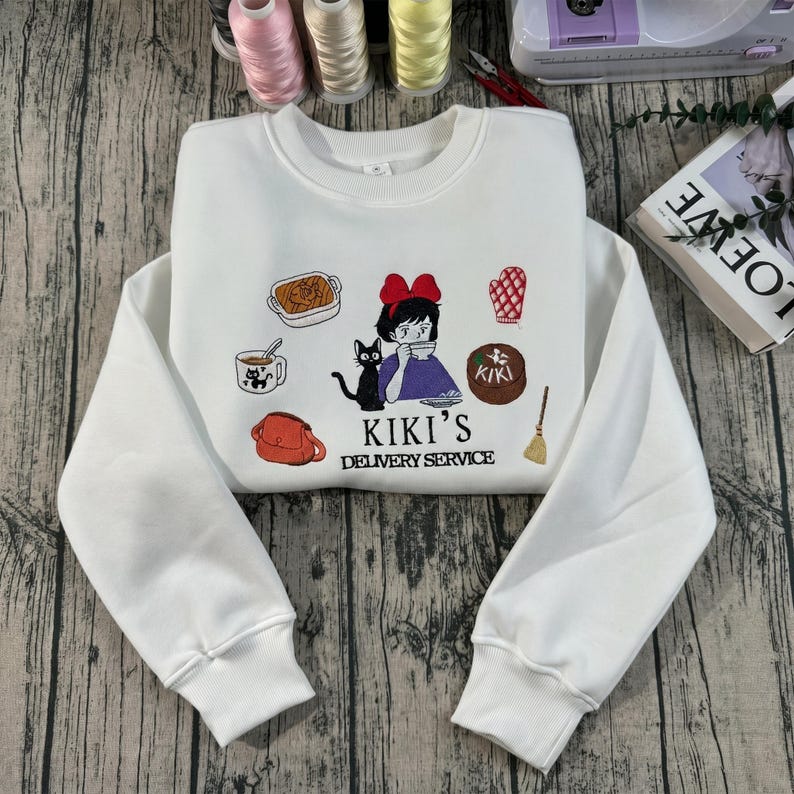 May include: White crewneck sweatshirt featuring a colorful embroidered design from Kiki's Delivery Service. The design includes a girl with a red bow, a black cat, and various delivery items. The text "KIKI'S DELIVERY SERVICE" is also embroidered.