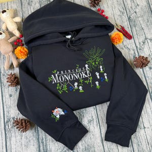 Pu&ograve; includere: Felpa con cappuccio nera con la scritta "PRINCESS MONONOKE" in un design verde e bianco. La felpa con cappuccio ha un cappuccio con coulisse e un disegno sulla manica. Il design include una creatura bianca e rossa e fiori blu.