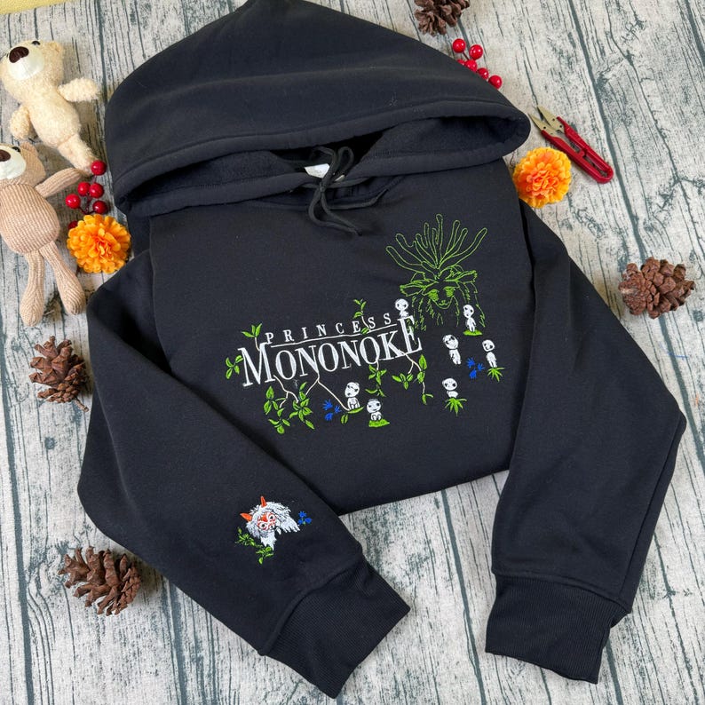 Pu&ograve; includere: Felpa con cappuccio nera con la scritta "PRINCESS MONONOKE" in un design verde e bianco. La felpa con cappuccio ha un cappuccio con coulisse e un disegno sulla manica. Il design include una creatura bianca e rossa e fiori blu.