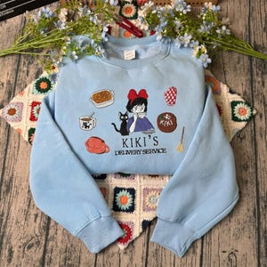 May include: A light blue sweatshirt featuring an embroidered design inspired by Kiki's Delivery Service. The design includes Kiki, a black cat, and various themed elements. The text "KIKI'S DELIVERY SERVICE" is also included.