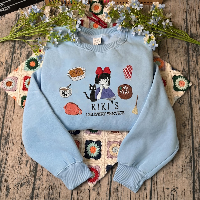 May include: A light blue sweatshirt featuring an embroidered design inspired by Kiki's Delivery Service. The design includes Kiki, a black cat, and various themed elements. The text "KIKI'S DELIVERY SERVICE" is also included.