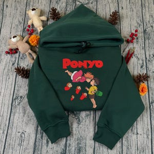 May include: A dark green hooded sweatshirt with the word "PONYO" in red. The sweatshirt features a cartoon graphic of two characters kissing, with red fish below. The garment is made of a soft material and has a drawstring.
