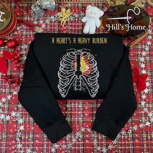 May include: Black long-sleeved sweatshirt featuring the text "A Heart's A Heavy Burden" above a graphic of a rib cage with a heart design. The heart is red and yellow. The sweatshirt is on a red and white plaid surface.