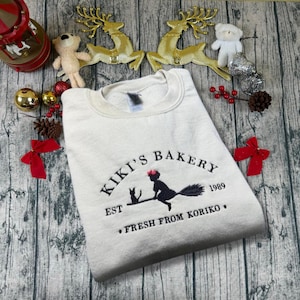 May include: Cream-colored sweatshirt with the embroidered text "Kiki's Bakery" and a graphic of a witch on a broom with a black cat. The text "EST 1989" and "FRESH FROM KORIKO" are also embroidered on the sweatshirt.