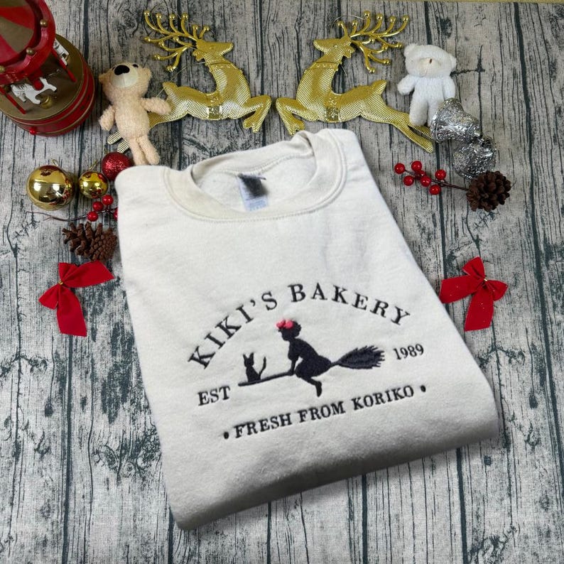 May include: Cream-colored sweatshirt with the embroidered text "Kiki's Bakery" and a graphic of a witch on a broom with a black cat. The text "EST 1989" and "FRESH FROM KORIKO" are also embroidered on the sweatshirt.