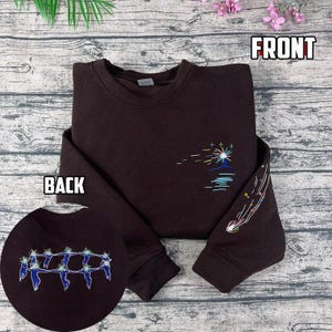 May include: A dark brown sweatshirt with embroidered celestial designs. The front features a sunburst over water, while the sleeve displays a shooting star. The back shows a circular embroidered design of figures. The word "FRONT" and "BACK" are printed on the image.
