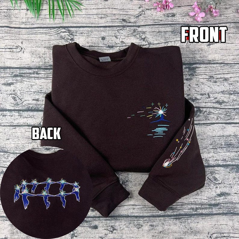 May include: A dark brown sweatshirt with embroidered celestial designs. The front features a sunburst over water, while the sleeve displays a shooting star. The back shows a circular embroidered design of figures. The word "FRONT" and "BACK" are printed on the image.