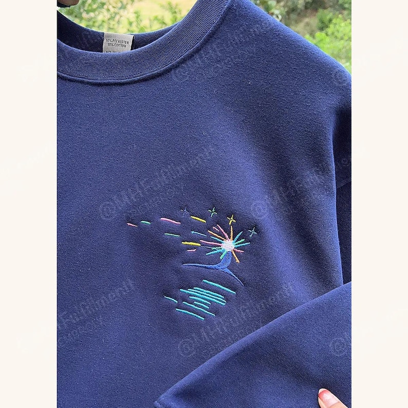 May include: Navy blue sweatshirt with an embroidered design of a colorful firework display over water. The embroidery features pink, yellow, green, and blue thread. The sweatshirt is made of a soft, comfortable material.
