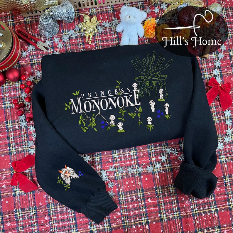 Pu&ograve; includere: Felpa nera con la scritta "Princess Mononoke" e un'illustrazione dello spirito della foresta e dei Kodama. La manica sinistra presenta un'immagine ricamata di una creatura simile a un lupo. Il design include accenti verdi, bianchi e blu.