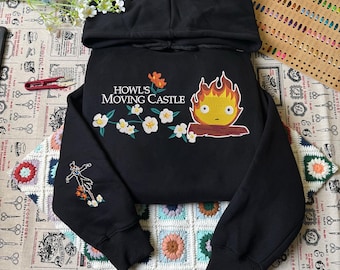 May include: A black hooded sweatshirt featuring a graphic of a fire character, white flowers, and the text Howl's Moving Castle. The sleeve displays an embroidered scarecrow figure with orange flowers. The hoodie has long sleeves and a hood.