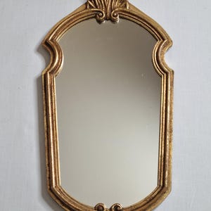 May include: A decorative gold-colored mirror with an ornate, arched frame. The mirror has a rectangular shape with a curved top and bottom. The frame features detailed scrollwork and a textured finish, adding a vintage aesthetic.
