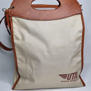 Może przedstawiać: Kremowa torba typu tote z brązowymi sk&oacute;rzanymi wykończeniami i uchwytem. Torba posiada logo UTA French Airlines w kolorze brązowym z przodu. Uchwyt ma owalne wycięcie do przenoszenia. Dołączony jest r&oacute;wnież pasek na ramię.