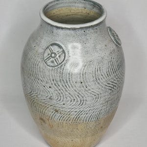 Puede incluir: Un jarrón de cerámica hecho a mano con un patrón texturizado en forma de onda. El jarrón es principalmente blanco con una base beige y dos diseños circulares en relieve. El interior es de color beige claro.