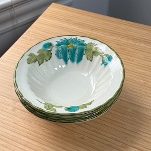 May include: A stack of four white ceramic bowls with a teal floral design and green trim. The bowls have a ridged interior and are stacked on a light brown wooden surface. The floral design includes teal flowers and green leaves.