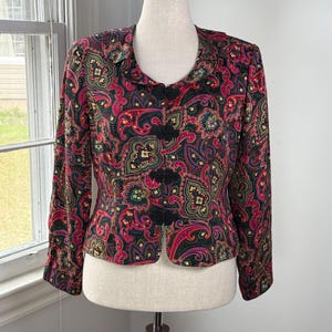 May include: A vintage paisley print jacket in shades of red, black, green, and gold. The jacket features a mandarin collar and three black frog closures. The long-sleeved jacket is displayed on a mannequin.