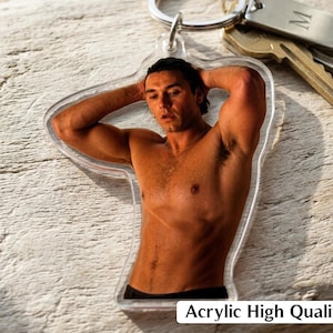 May include: A clear acrylic keychain featuring a photographic print of a shirtless person with their hands behind their head. The keychain includes a silver-toned metal ring and a rectangular metal tag engraved with the letter M. The text Acrylic High Quality is visible.
