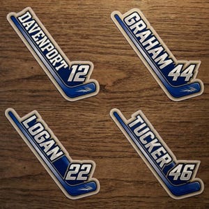 Könnte beinhalten: Vier Hockey-Stick-Aufkleber mit Namen und Nummern: Davenport 12, Graham 44, Logan 22 und Tucker 46. Jeder Aufkleber ist blau und weiß, in Form eines Hockeyschlägers, mit weißem Rand, vor einem Holzmaserungshintergrund.