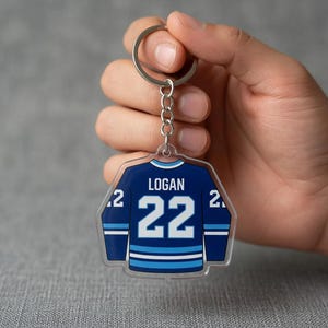 May include: A clear acrylic hockey jersey-shaped keyring. The navy blue jersey features white and light blue stripes and the number 22. The name "LOGAN" is printed above the number.
