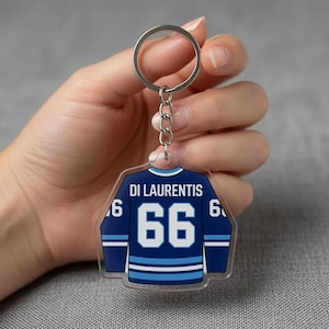 May include: A clear acrylic hockey jersey keyring. The navy blue jersey has white and light blue details, with the name "DI LAURENTIS" and the number "66". It is attached to a silver metal ring for keys.
