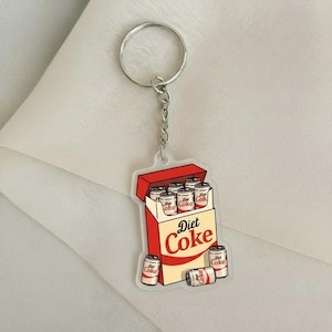 Puede incluir: Un llavero de Diet Coke con una caja roja y beige con la inscripción "Diet Coke". La caja está abierta, revelando varias latas pequeñas de Diet Coke. El llavero tiene una cadena y un anillo plateados.