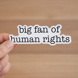 May include: A white sticker with the text "big fan of human rights" in black, held by a hand. The sticker has a unique shape and is set against a blurred wooden background.