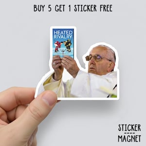 May include: A sticker featuring an image of a hockey game with the text "Heated Rivalry" held by a person. The sticker is held in front of a person wearing glasses and a white robe. The text "Buy 5 Get 1 Sticker Free" is at the top.