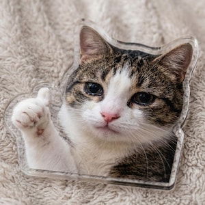 May include: A white ceramic mug with a black handle and the text "But First, Coffee" in a bold, black font. The mug is set against a blurred background. The mug appears to be approximately 10 cm tall.