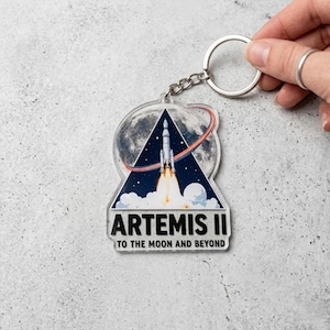 May include: Clear acrylic keychain featuring a rocket launching from a triangular design with the text "ARTEMIS II TO THE MOON AND BEYOND." The keychain has a silver chain and ring.