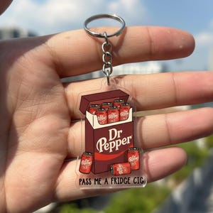 Peut inclure: Un porte-clés en acrylique transparent avec un design sur le thème de Dr Pepper. Le porte-clés représente un paquet de canettes de Dr Pepper avec le texte "Dr Pepper" et "PASS ME A FRIDGE CIG". Il est attaché à une chaîne et un anneau en métal argenté.