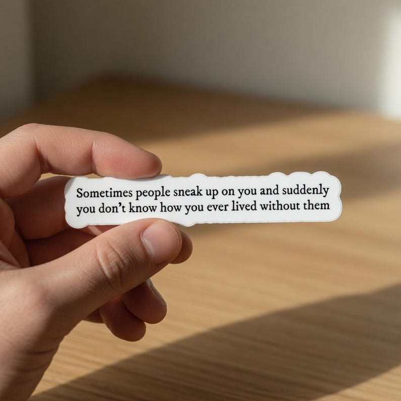 May include: A white sticker with a wavy edge, held by a hand. The sticker has black text that reads, "Sometimes people sneak up on you and suddenly you don't know how you ever lived without them."