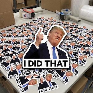 Peut inclure: Une collection d'autocollants blancs avec un portrait d'un homme en costume et cravate, avec le texte "I DID THAT". Les autocollants sont éparpillés sur une table, avec du matériel d'emballage en arrière-plan.