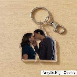 May include: A clear acrylic keychain featuring a printed image of a person with dark hair wearing a dark blue shirt kissing another person with dark hair wearing a dark gray long-sleeve shirt. The keychain includes a silver metal ring and a lobster clasp.