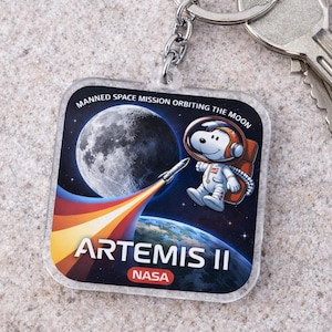 May include: Clear square keychain with a Snoopy astronaut design, a rocket, and the moon. The keychain has the text "Artemis II NASA" and "Manned Space Mission Orbiting the Moon."